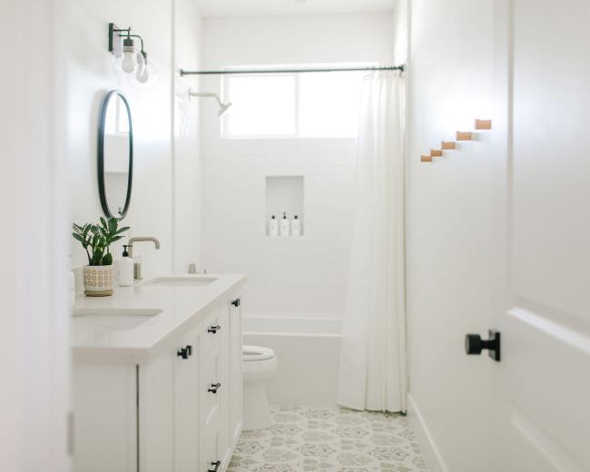 The image shows a bright, modern bathroom featuring a white vanity, a circular mirror, a bathtub with a shower curtain, and decorative wall hooks.