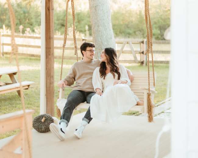 A couple sits together on a swing under a porch, surrounded by greenery and wooden fencing.