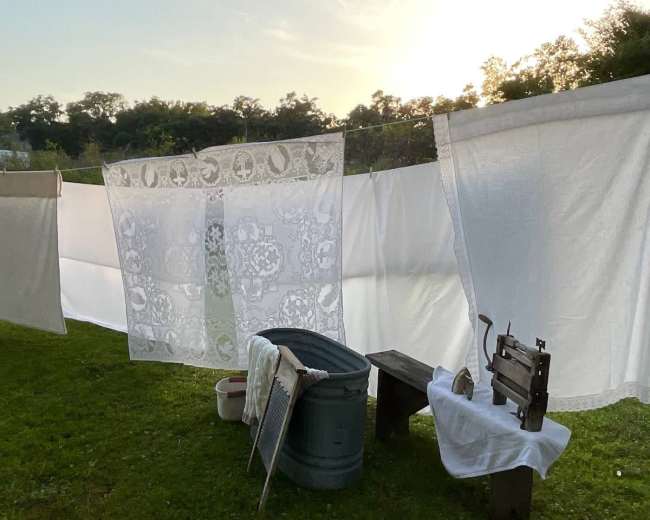 Clean laundry hangs on a clothesline in a grassy area, with a vintage wash tub and ironing board nearby, illuminated by the soft light of sunset.