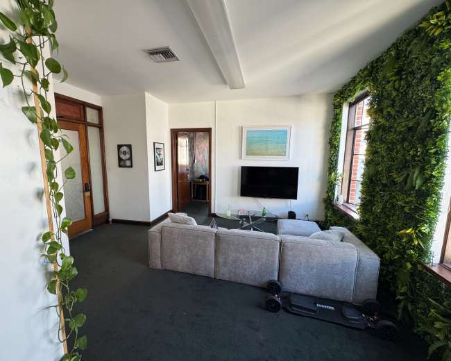 The image shows a modern living room with a grey sectional sofa, a coffee table, and a television, surrounded by decorative plants on the walls.