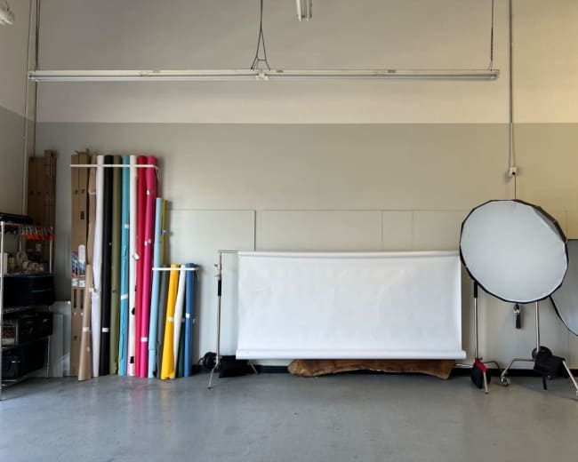 The image shows a spacious studio with rolls of colored backdrop materials on the left and photography equipment, including two reflectors, on the right.