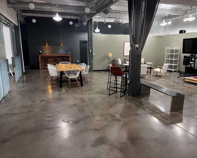 The image shows a spacious, minimalist interior with a mix of seating options, including a dining table, chairs, and couches, on a polished concrete floor.