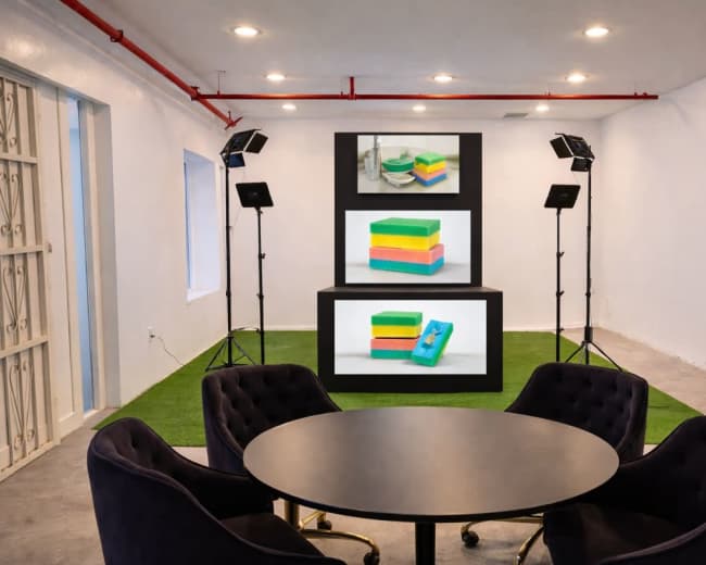 The image shows a bright, minimalist room with a round black table and plush chairs, featuring three stacked screens displaying colorful sponges.