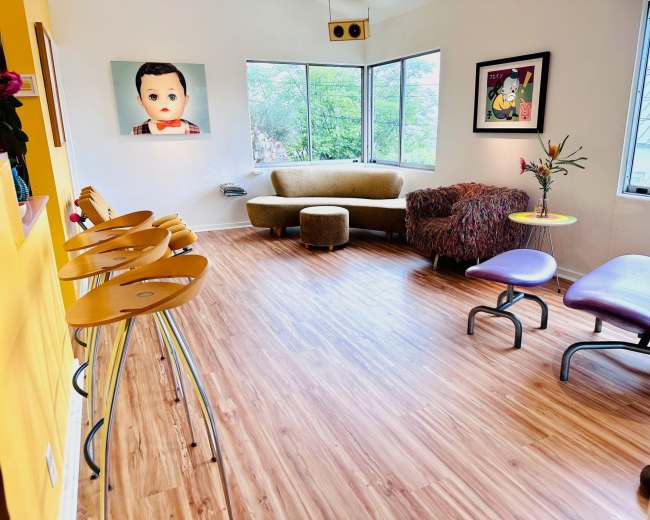 A brightly lit room featuring a cozy seating area with a curved couch and a colorful chair, along with a row of wooden bar stools against a yellow wall.