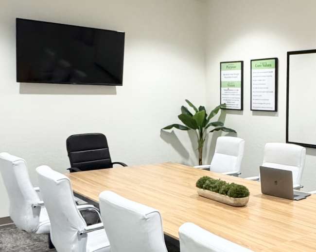 A modern conference room features a large wooden table surrounded by white chairs, a laptop on the table, and a wall-mounted TV.