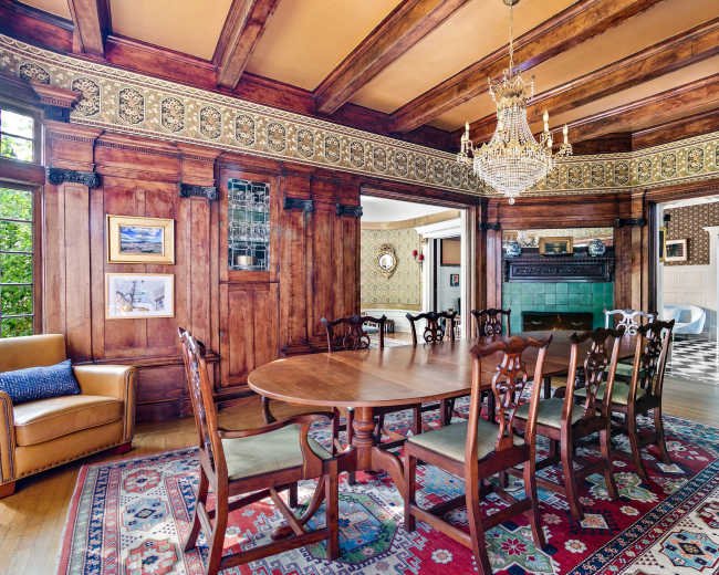 The image shows an ornate dining room with dark wood paneling, a chandelier, and a large wooden table surrounded by chairs, all complemented by a patterned rug and wallpaper.