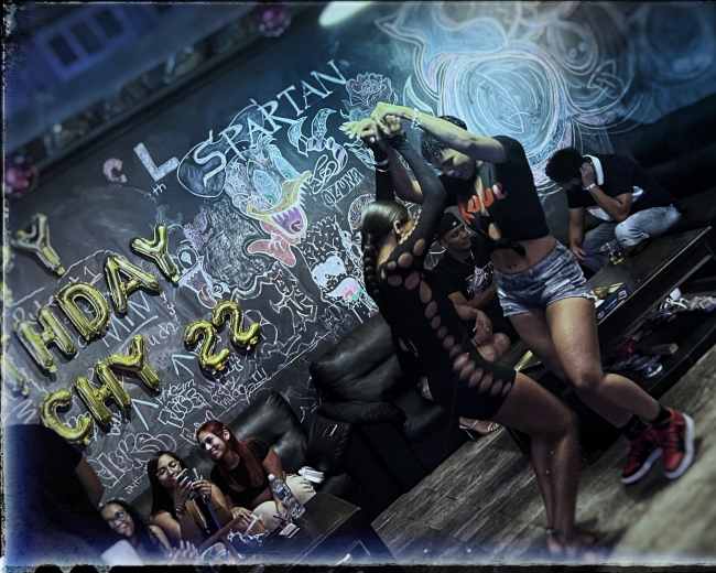 Two dancers perform in a lively venue decorated with colorful balloons and graffiti on the walls while several onlookers enjoy the scene from nearby tables.