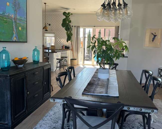 The image shows a modern dining area featuring a long black table surrounded by chairs, a large green plant in the center, and a bright open kitchen visible in the background.