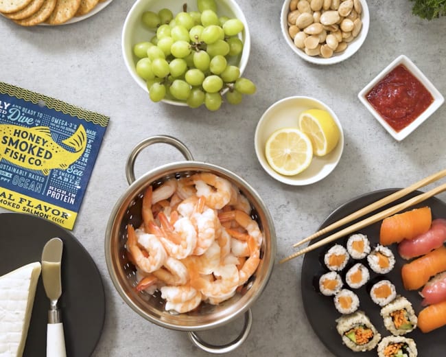 A variety of food items including shrimp in a colander, sushi rolls, crackers, cucumbers, grapes, nuts, lemon slices, and a bowl of sauce, arranged on a table.