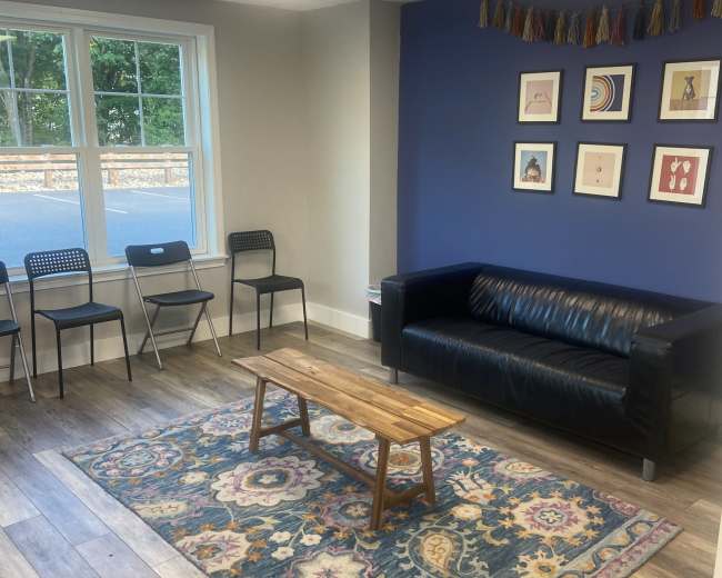 The image shows a modern lounge area with a blue accent wall, a black sofa, a wooden coffee table, patterned rug, and some chairs arranged around the space.