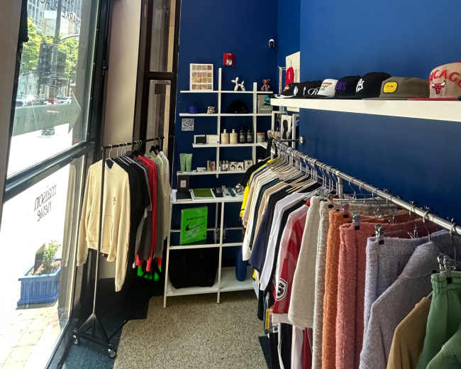 The interior of a retail store features a clothing rack filled with various shirts and sweatshirts, alongside shelves displaying hats and small merchandise against a blue wall.