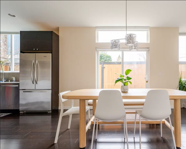 The image shows a modern dining area with a wooden table, white chairs, and a potted plant, overlooking a patio through large windows.