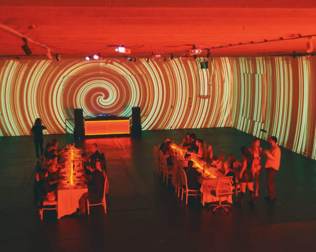 A group of people is gathered around a long table in a dimly lit room with swirling red and orange light projections on the walls.