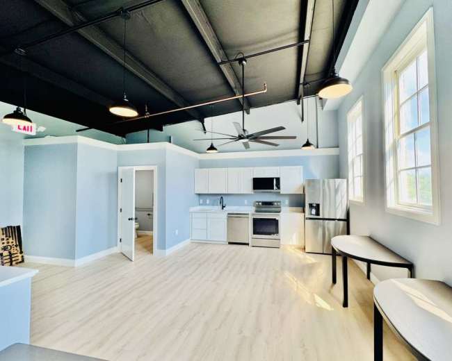 The image shows a modern, open-concept kitchen and living area with light blue walls, stainless steel appliances, and large windows.