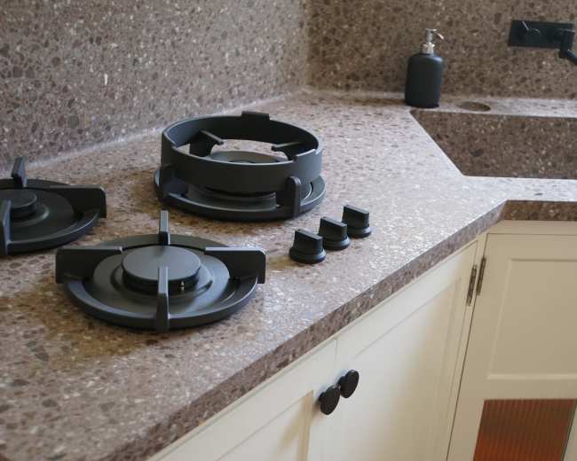 The image shows a modern kitchen countertop made of speckled stone with gas burners and accessory components arranged on it.