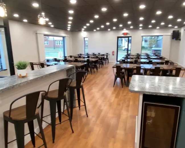 The image shows an interior view of a restaurant with rows of tables and chairs, a bar counter, and several ceiling lights illuminating the space.