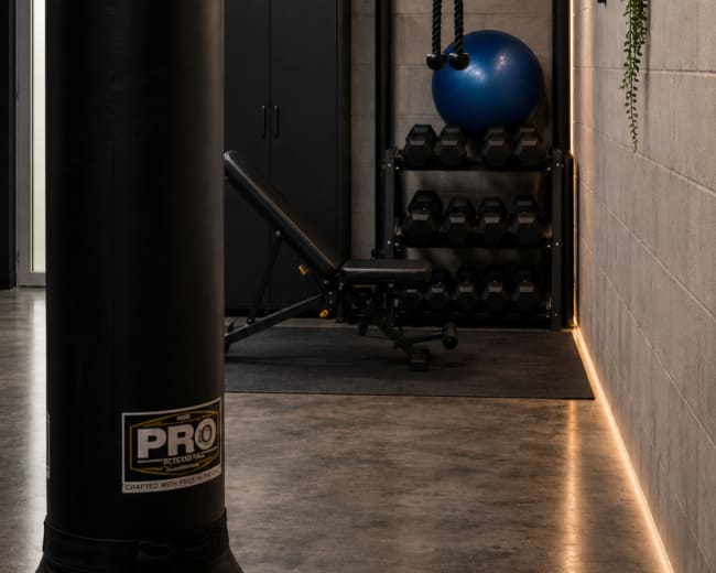 The image shows a modern fitness room equipped with a punching bag, dumbbells, a blue exercise ball, and a weight bench against a gray wall, illuminated by geometric ceiling lights.
