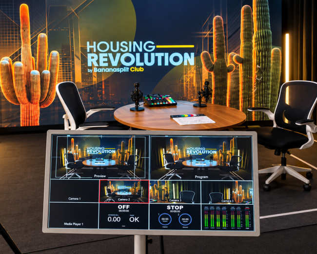 The image shows a modern studio setup for a show titled "Housing Revolution," featuring a large screen with graphics, a round table, two chairs, and production equipment including cameras and a control monitor.