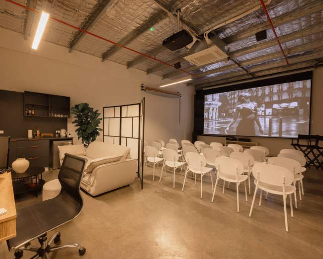 The image shows a modern meeting room with a projection screen at the front, arranged seating in white chairs, a couch, and a small kitchenette area.