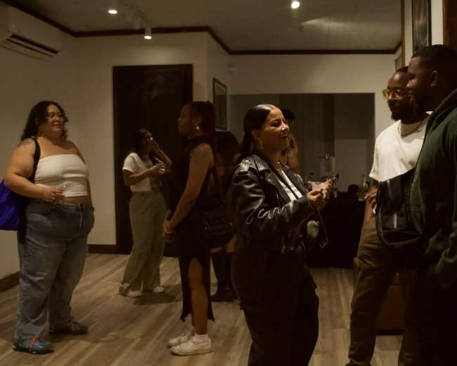 A group of people converse in an indoor space with wooden flooring and art on the walls.