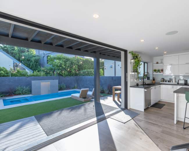 The image shows a modern kitchen with an open layout that leads to a backyard featuring a small pool and lawn area.