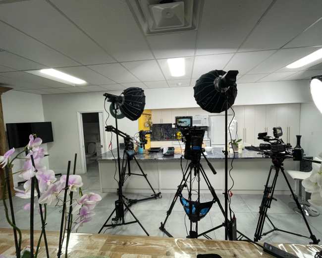 A well-lit kitchen set up for filming, featuring multiple cameras and lighting equipment in front of a counter.