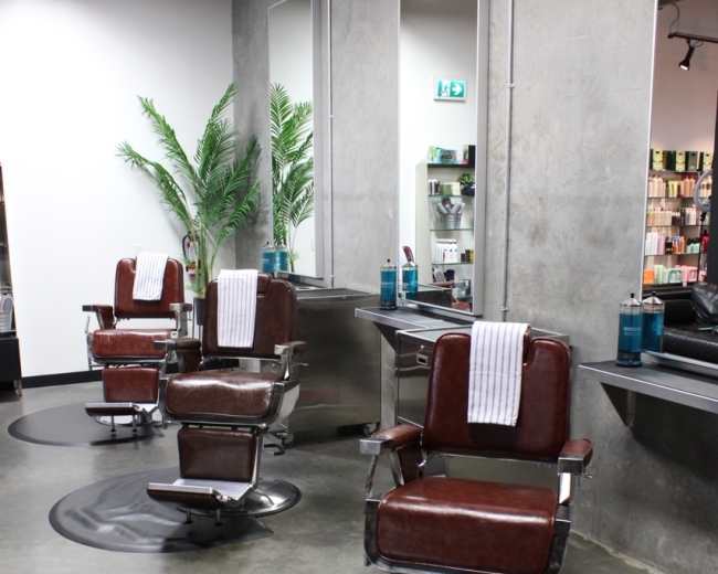 The image shows a modern barbershop with three vintage-style barber chairs arranged in front of large mirrors and a concrete wall.