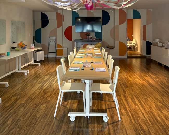 A long table is set with colorful materials in a modern meeting room featuring abstract wall decorations and streaming lights overhead.