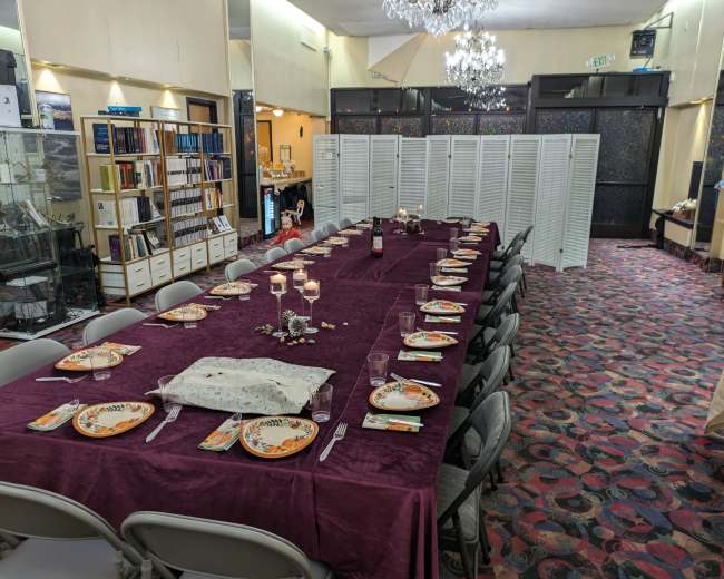 A long dining table is set with plates and glassware, surrounded by chairs, in a banquet room adorned with patterned carpeting and a chandelier.