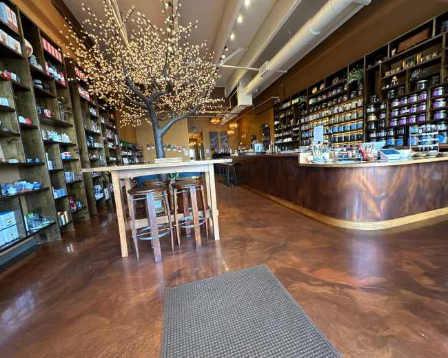A brightly lit shop features wooden shelves filled with various products, a central table with chairs, and a decorative tree with lights.