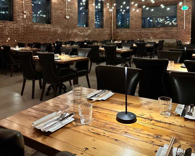 The image shows a spacious restaurant interior with exposed brick walls, wooden tables set for dining, and warm overhead lighting.
