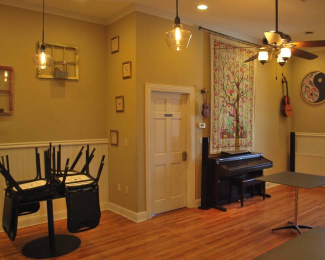 The interior of a cozy room features a piano against one wall, a table, and stacks of chairs upside down on another.