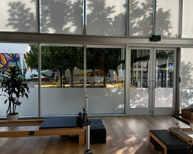 The image shows a modern fitness studio with large glass doors and windows, revealing an outdoor area with trees and colorful murals.