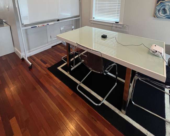 The image shows a conference room with a large white table, transparent chairs, and a whiteboard on a stand against a light-colored wall.