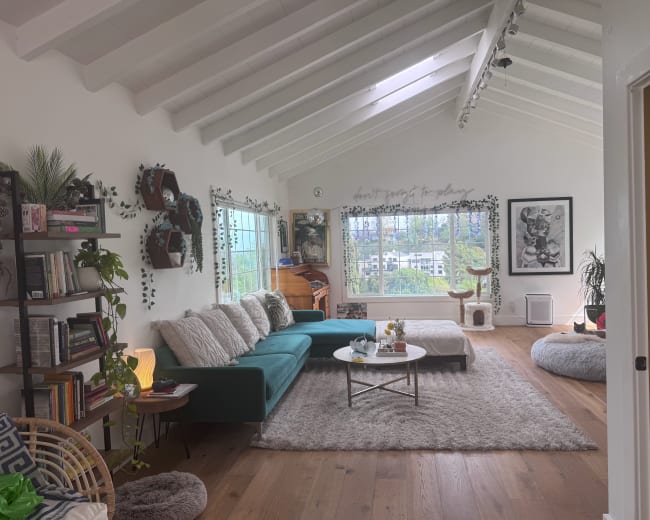 The living room features a teal couch, a large rug, and panoramic windows with greenery outside, along with shelves filled with books and decorative plants.