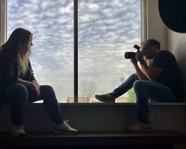 A woman sits on a windowsill while a man with a camera photographs her from a nearby bench.
