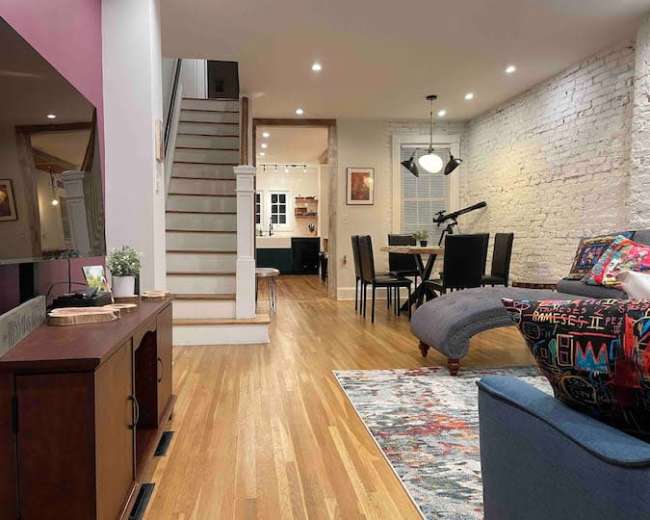 The image shows a modern living space with hardwood floors, featuring a staircase, a dining area, and a cozy seating arrangement.