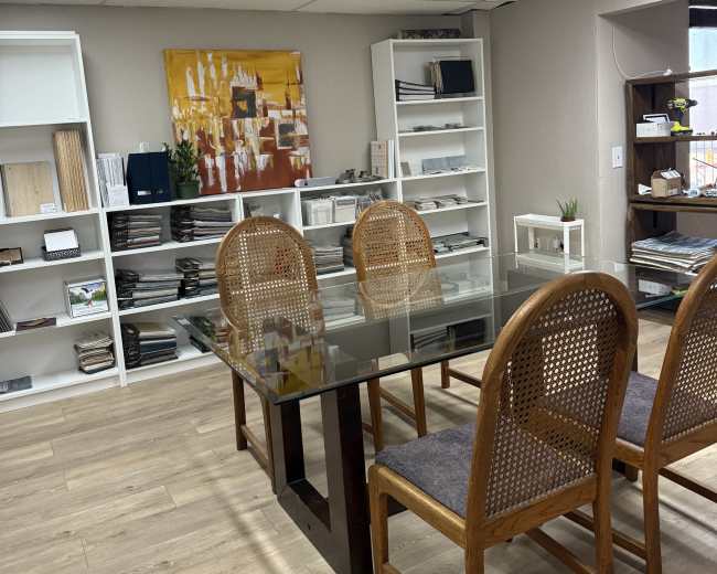 The image shows a well-lit office space with a glass meeting table surrounded by four wooden chairs, complemented by shelves filled with various materials and decor.
