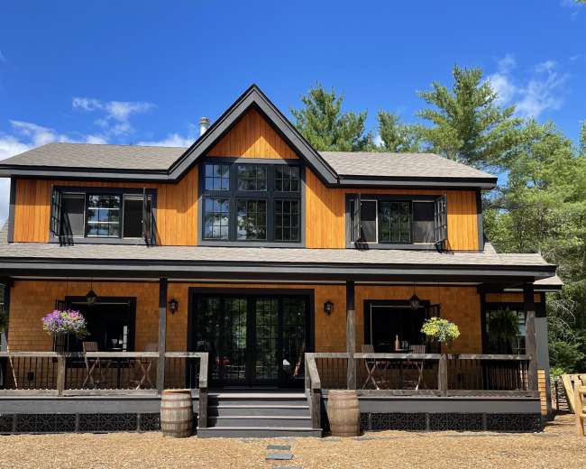 A two-story house with a wooden exterior, large windows, and a covered porch, surrounded by trees and gravel.
