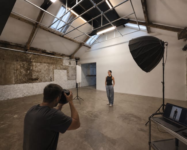 A photographer is taking a portrait of a model in a spacious, well-lit studio with exposed beams and large windows.