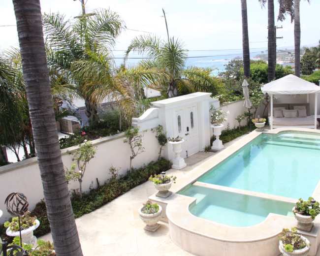 A residential backyard with a swimming pool surrounded by manicured plants and palm trees, overlooking a coastal view.