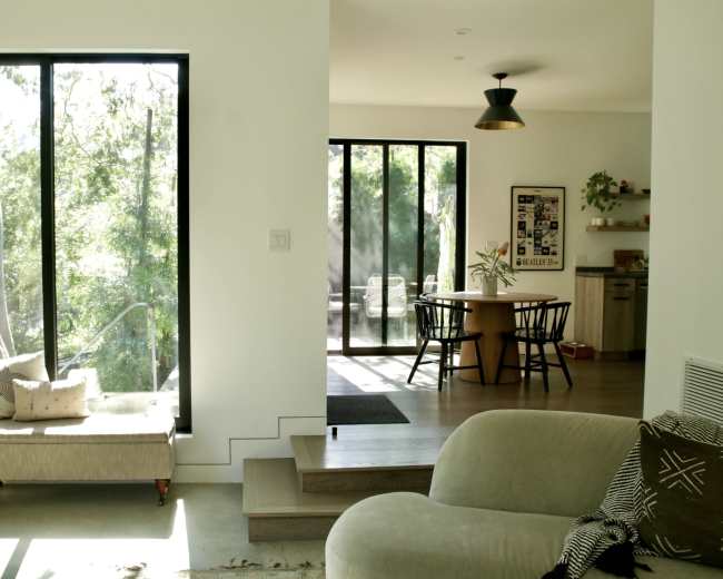 The image features a modern living space with large windows allowing natural light to illuminate the room, furniture arranged for comfort, and a dining area visible in the background.
