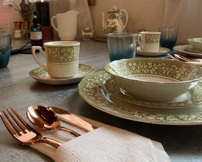 A dining table set with green and white china, silver utensils, and glasses, complemented by soft natural light coming through the curtains.