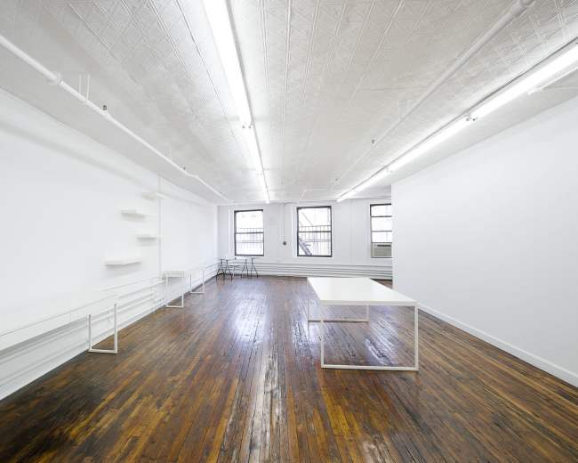 The image shows a spacious, minimalistic room with a hardwood floor, white walls, several empty shelves, and a large white table at the center.
