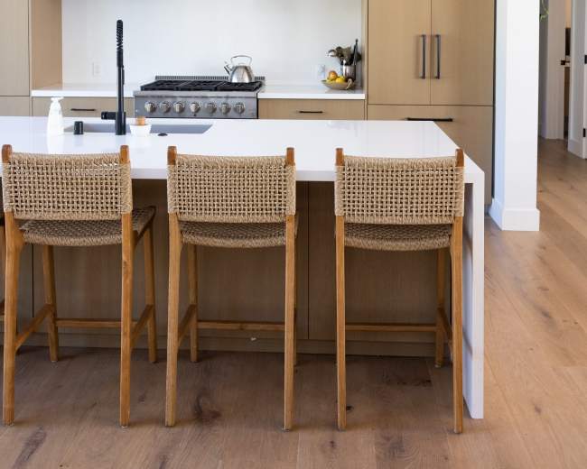 A modern kitchen features a large island with three wooden bar stools, a black pendant light overhead, and beige cabinetry.