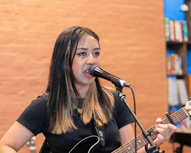 A woman sings into a microphone while playing an acoustic guitar during a performance.