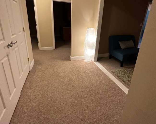 A well-lit hallway features beige walls, carpeted flooring, and a sleek lamp beside an open doorway leading to a small, furnished room.