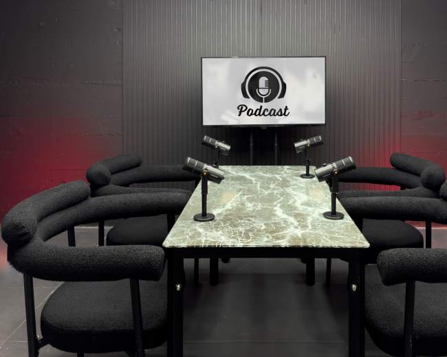 A modern podcast studio features a marble table surrounded by black chairs, with multiple microphones positioned on stands, and a screen displaying the word "Podcast."