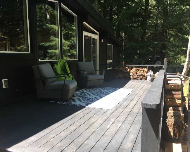 A deck with two chairs and a patterned rug is situated in a wooded area, alongside a stacked pile of firewood.