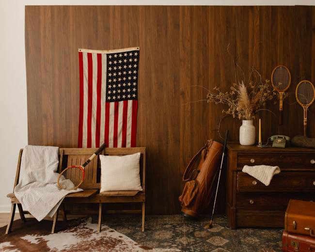 The image features a rustic indoor scene with a wooden wall, an American flag, a vintage tennis racket, a cream-colored vase with dried flowers, a wooden dresser, and various sports equipment arranged artistically.